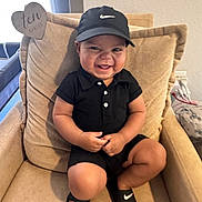Devin is registered to the contest to win money with this photo: toddler, child, smiling, black_clothing, nike_cap, nike_slides, armchair, indoor, happy, cute, portrait, sitting, baby, footwear, foot, furniture, person, human, cushion, home