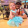 baby, child, man, glasses, cap, dress, blue_dress, tongue_out, smile, indoor, restaurant, table, chair, lights, ceiling, people, casual, person, holding, happy