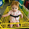 baby, child, shopping_cart, yellow_cart, red_hair, pink_dress, cherry_pattern, face, eyes, shopping, store, retail, seatbelt, floor, plastic_cooler, boxes, shelves, indoor, person, expression