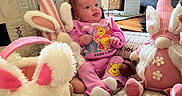 Elena is registered to the contest to win money with this photo: baby, blanket, bunny_ears, child, cute, decor, gnome_doll, indoors, lamp, living_room, pajamas, pink_clothing, plush_toy, quilt, seated, soft_toys, stuffed_animal, tissue_box, wall_art, wooden_table