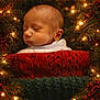 Isaac is registered to the contest to win money with this photo: baby, infant, sleeping, knitwear, red_sweater, green_blanket, christmas_lights, pine_branches, berries, holiday, festive, cozy, portrait, closeup, warm_lighting, soft_focus, indoors, cute, peaceful, celebration