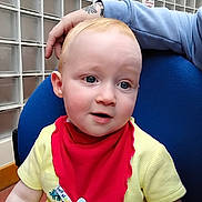 Eli is registered to the contest to win money with this photo: adult_hand, baby, blue_eyes, chair, child, close_up, curious_expression, cute, face, glass_blocks, indoor, infant, person, poster, red_bib, seated, skin, tattoo, yellow_clothing, young
