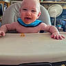 baby, infant, child, high_chair, smiling, tongue_out, portrait, indoor, home, feeding, food, tray, blue_bib, happy, sitting, face, hands, fan, television, rustic_interior