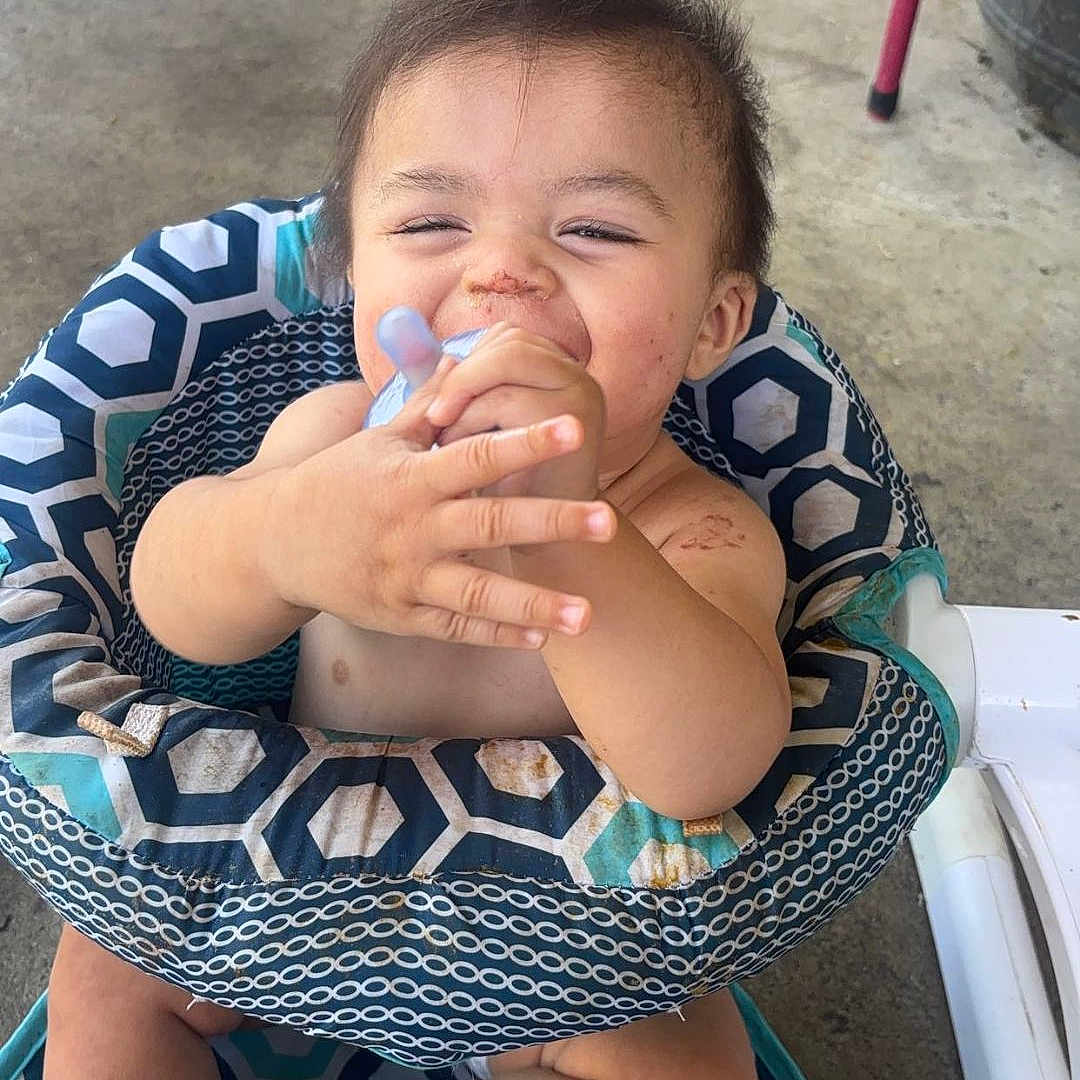 Houston joined the competition — help win amazing prizes! baby, bare_chest, barefoot, bottle, child, concrete_floor, cute, dirty_face, feeding, happy, high_chair, indoor, infant, patterned_fabric, person, playful, sitting, smiling, toys, young