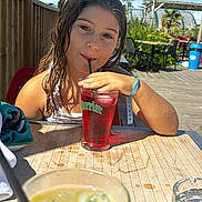Alice participe au concours pour gagner de l'argent avec cette photo : child, girl, drink, straw, glass, table, outdoor, sunny, daylight, wooden_table, beverage, wet_hair, red_drink, yellow_drink, ashtray, greenery, chair, sky, clouds, relaxation