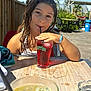 child, girl, drink, straw, glass, table, outdoor, sunny, daylight, wooden_table, beverage, wet_hair, red_drink, yellow_drink, ashtray, greenery, chair, sky, clouds, relaxation