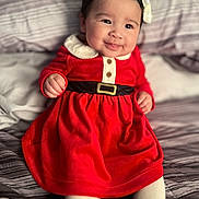 Kailani is registered to the contest to win money with this photo: baby, child, smiling, red_dress, white_bow, bed, striped_bedding, infant, cute, indoor, portrait, happy, clothing, person, sitting, small_hands, face, head, cozy, soft