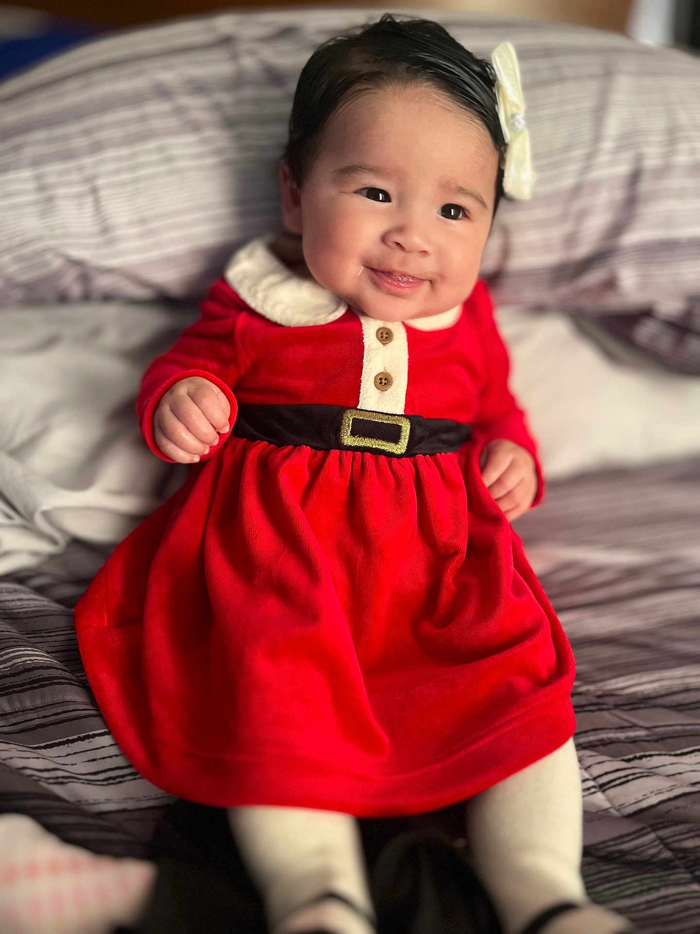 Kailani is registered to the contest to win money with this photo: baby, child, smiling, red_dress, white_bow, bed, striped_bedding, infant, cute, indoor, portrait, happy, clothing, person, sitting, small_hands, face, head, cozy, soft