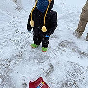 Isaac is registered to the contest to win money with this photo: child, snow, winter_clothing, yellow_hat, face_mask, green_slippers, snow_shovel, snowy_ground, winter, outdoor, cold_weather, black_jacket, gloves, person, footwear, snowfall, fence, pants, standing, looking_up