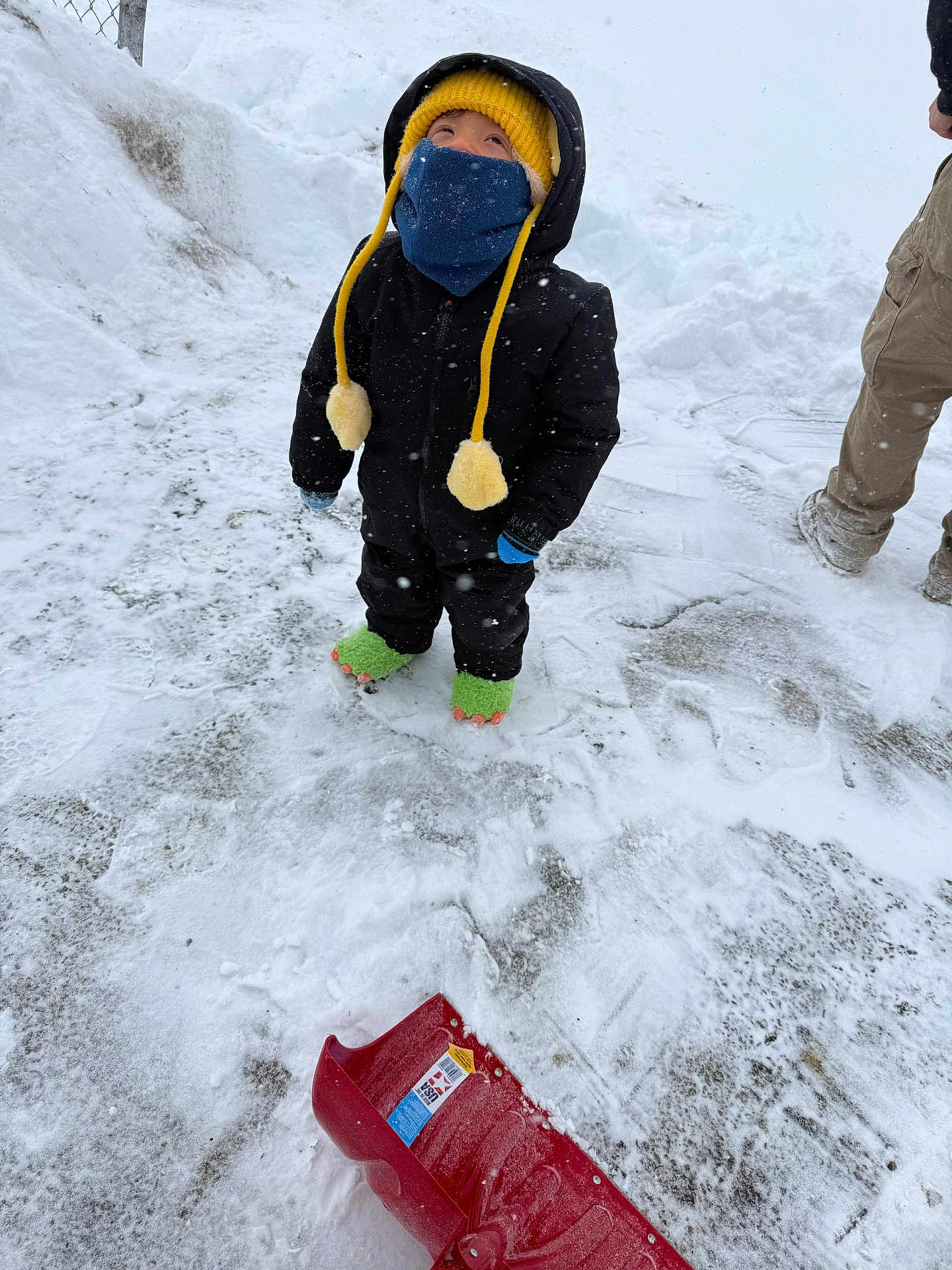Isaac is registered to the contest to win money with this photo: child, snow, winter_clothing, yellow_hat, face_mask, green_slippers, snow_shovel, snowy_ground, winter, outdoor, cold_weather, black_jacket, gloves, person, footwear, snowfall, fence, pants, standing, looking_up