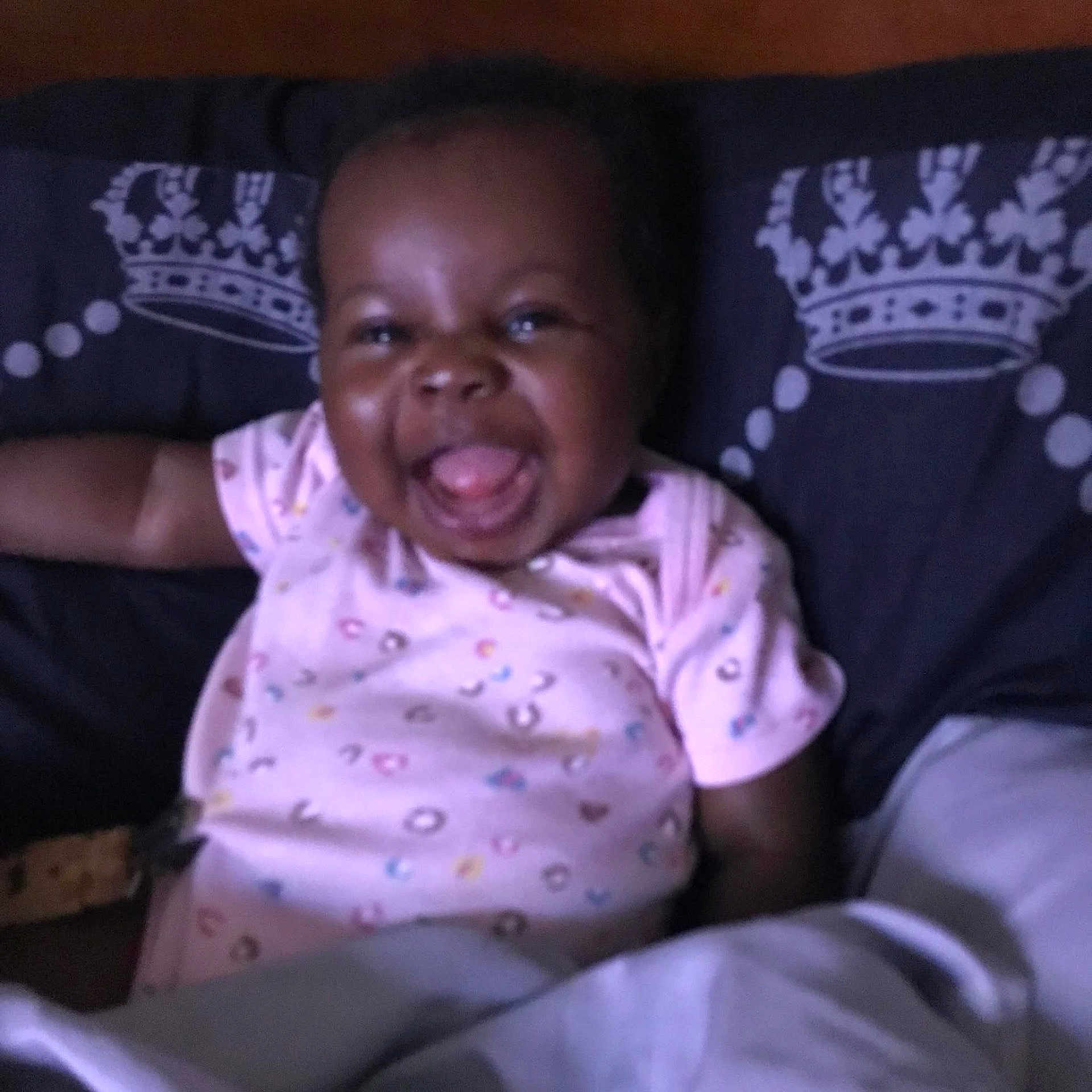 Dahveyah is registered to the contest to win money with this photo: baby, child, smiling, tongue_out, pink_clothing, onesie, pillow, crown_pattern, blanket, indoor, happy, cute, infant, face, person, lying_down, dark_background, closeup, young_child, portrait