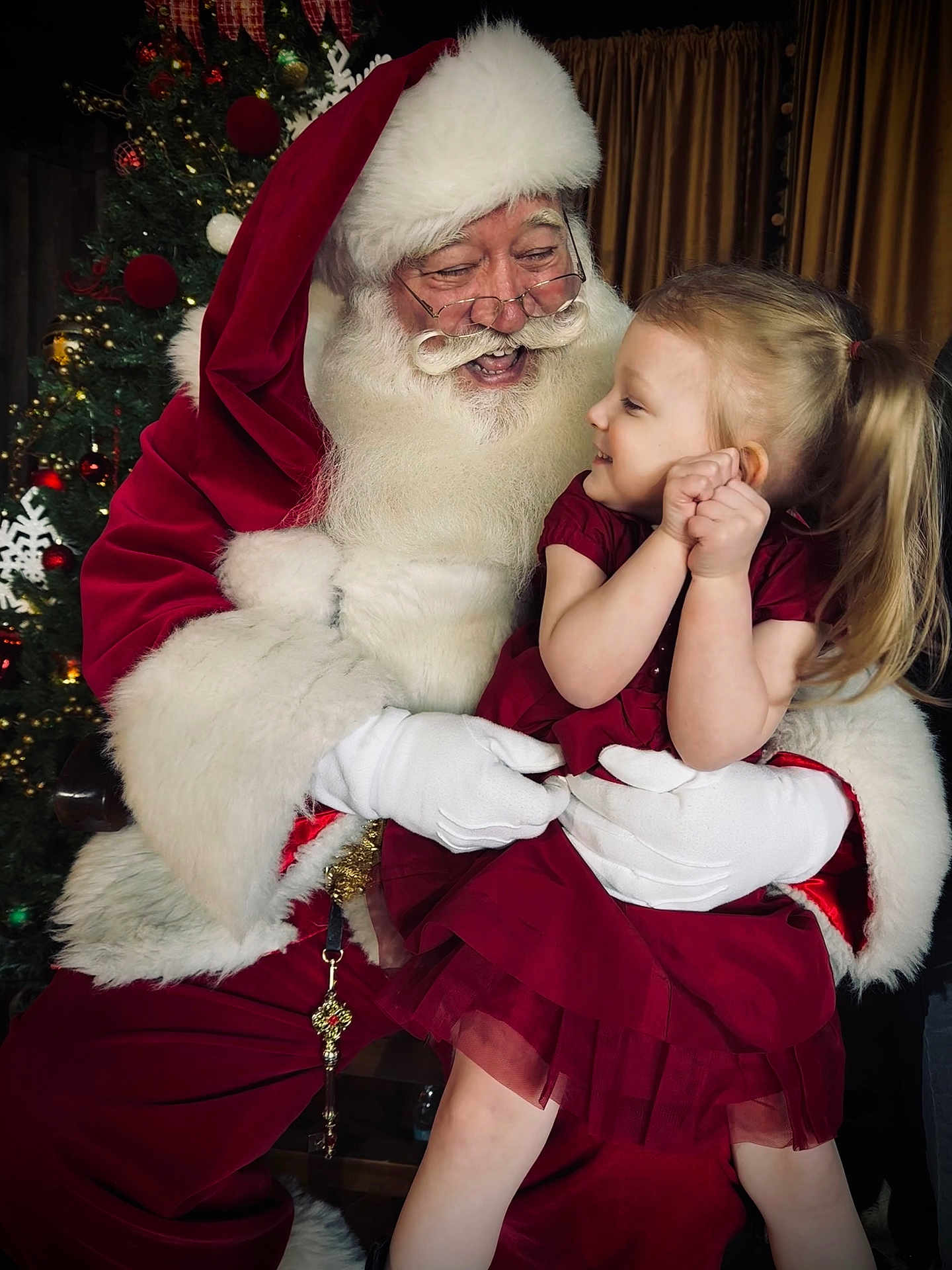 Mae is registered to the contest to win money with this photo: child, santa_claus, christmas_tree, holiday, festive, red_dress, white_beard, glasses, gloves, smiling, decorations, celebration, indoor, portrait, happy, toddler, christmas_lights, seasonal, family, joy