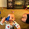 baby, child, toy, indoor, carpet, fireplace, furniture, person, onesie, hand, face, smile, adult, legs, living_room, sitting, play, curious, star_pattern, blue_toy