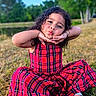 child, girl, red_dress, plaid, outdoor, grass, smiling, posing, sitting, playful, curly_hair, nature, daylight, footwear, sandals, greenery, fun, portrait, cute, happy