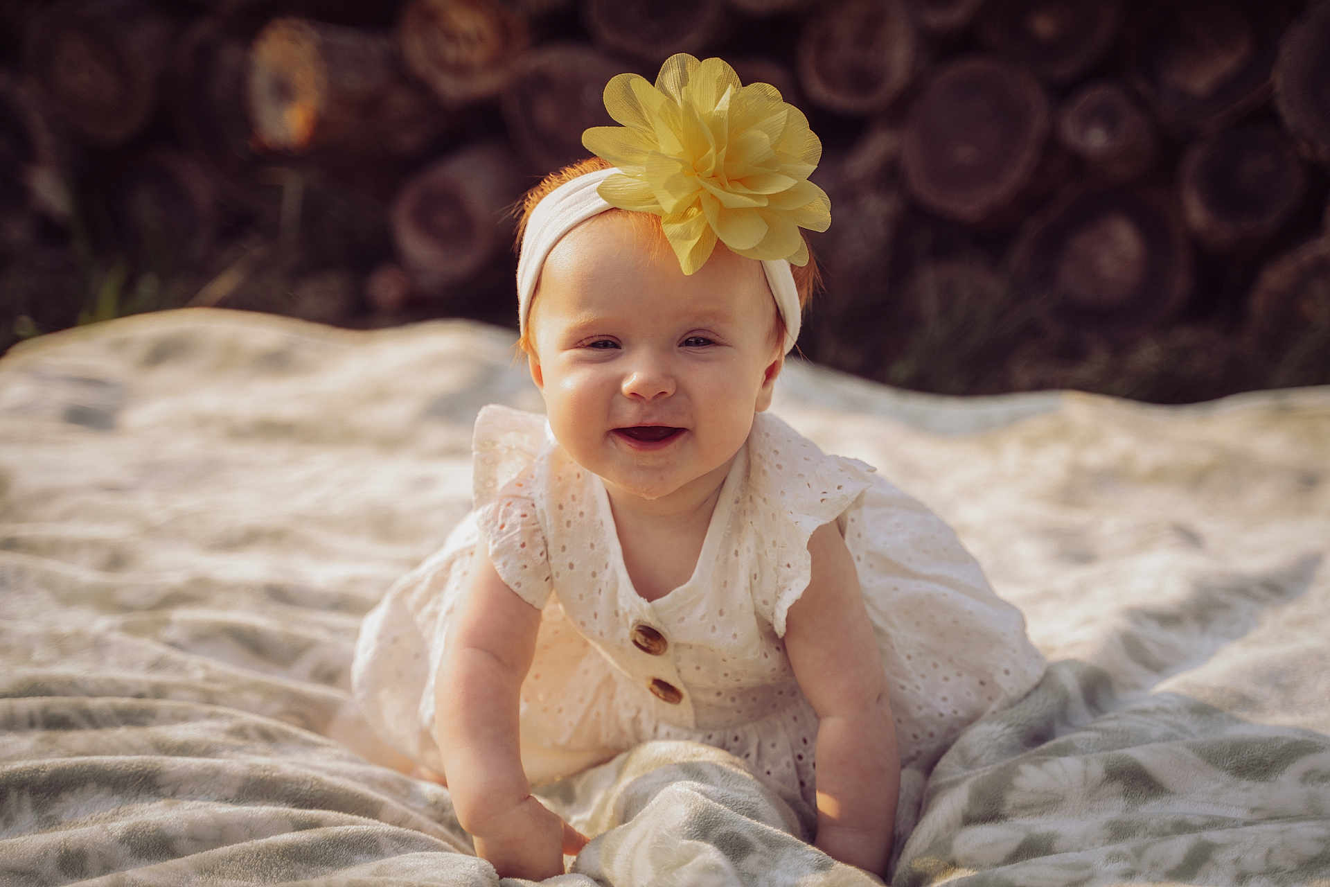 Harper is registered to the contest to win money with this photo: baby, child, crawling, flower_headband, yellow_flower, white_dress, blanket, outdoor, smiling, cute, infant, headband, nature, sunlight, wood_logs, portrait, happy, young_child, soft_texture, innocence