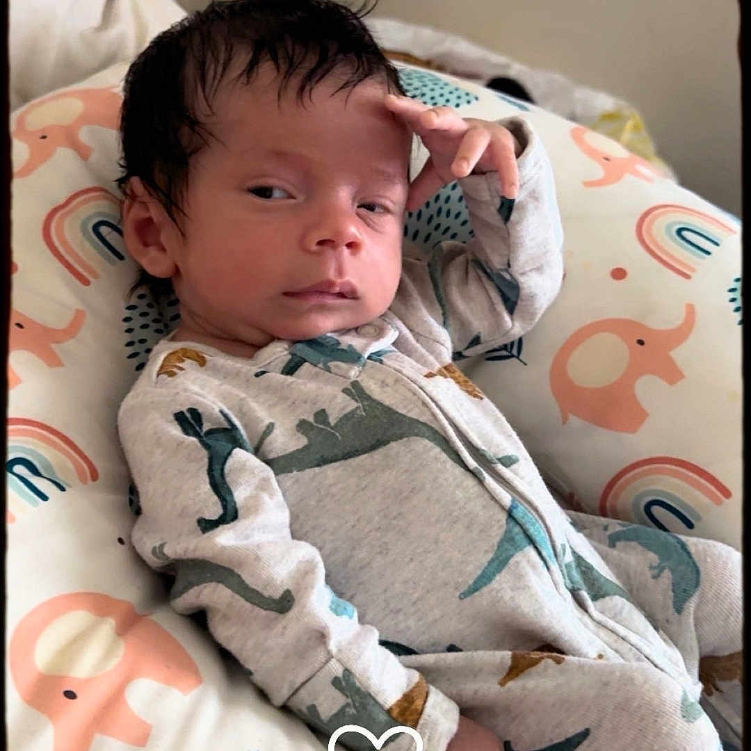 Halim is registered to the contest to win money with this photo: baby, child, cozy, cute, dinosaur_pattern, elephant_pattern, expression, face, hand_on_forehead, indoors, infant, onesie, person, pillow, portrait, rainbow_pattern, resting, sleepwear, soft_lighting, young_child