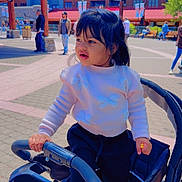 Divgun is registered to the contest to win money with this photo: toddler, stroller, outdoor, child, curious, white_sweater, dark_hair, people, plaza, buildings, daylight, casual_clothing, urban, walking, bench, tree, pavement, blue_sky, sunny, family