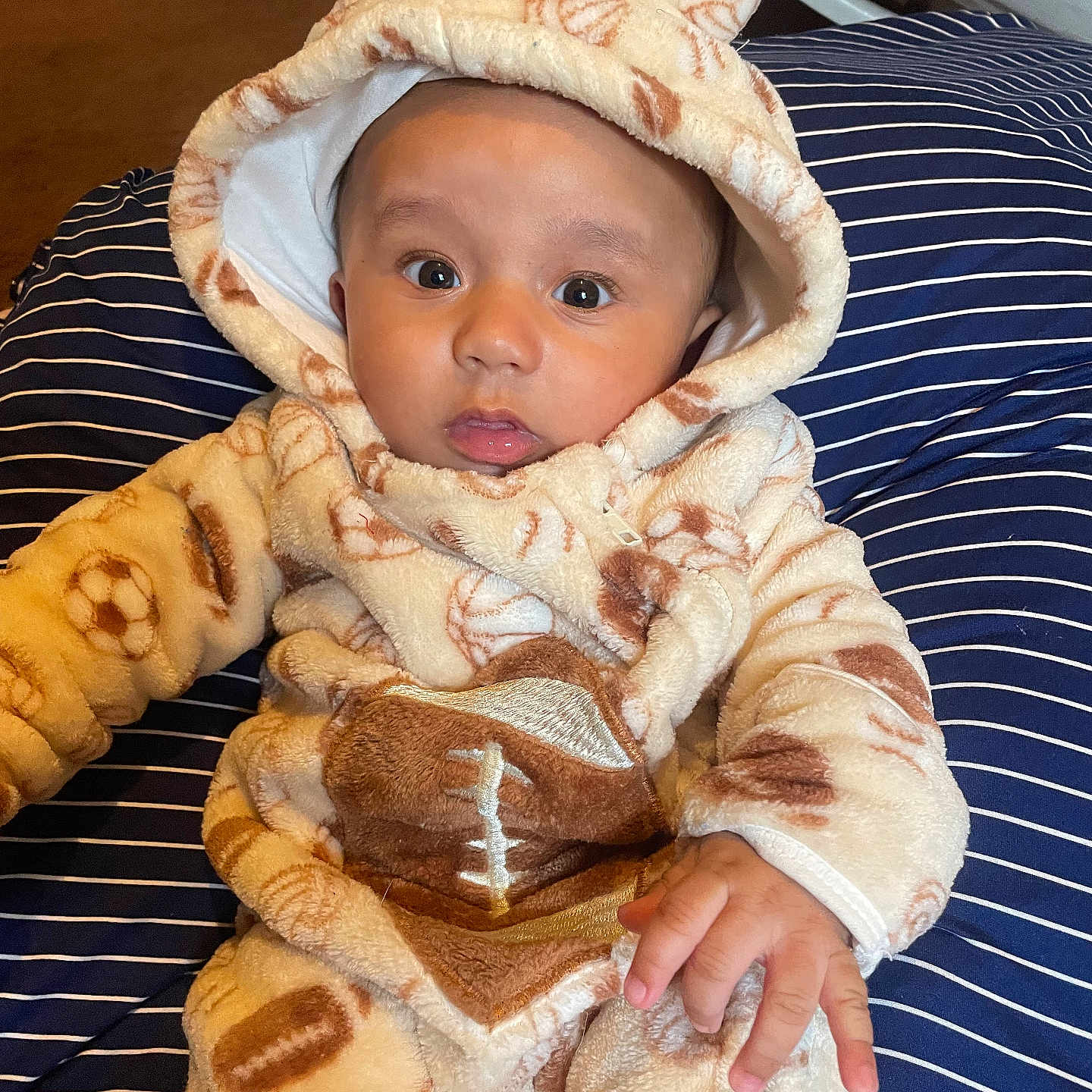 Kirk Jr is registered to the contest to win money with this photo: baby, infant, child, fleece_suit, sports_theme, football_pattern, hooded_clothing, striped_pillow, indoor, carpet, curious_expression, sitting, hand, face, eyes, cozy, warm_clothing, cute, portrait, person