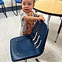 Zy'Aire is registered to the contest to win money with this photo: toddler, child, chair, blue_chair, floor, table, wooden_table, curly_hair, cartoon_shirt, indoor, tile_floor, waiting_room, classroom, playful, expression, young_child, person, furniture, standing, casual_clothing