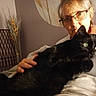 person, cat, black_cat, glasses, indoor, holding, pet, green_eyes, elderly, cozy, warm_lighting, furniture, decor, dried_plants, portrait, animal, companion, home, resting, closeup