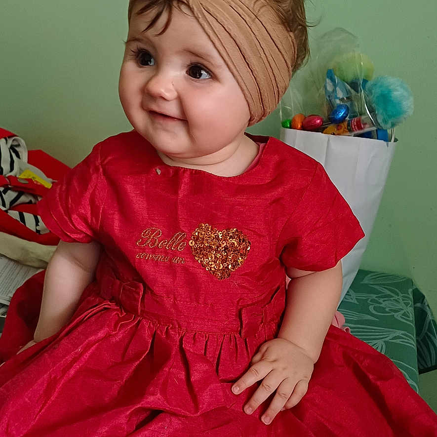 Anaïs a rejoint le concours — aidez-le/la à gagner de superbes lots ! baby, child, clothing, cute, face, foot, gift_bag, green_wall, hand, happy, headband, indoor, patterned_surface, portrait, red_dress, shells, sitting, smiling, toys, young