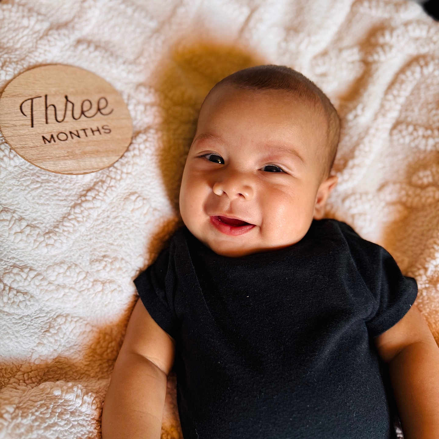 Noah is registered to the contest to win money with this photo: baby, black_onesie, blanket, cute, face, happy, indoors, infant, lying_down, milestone, person, portrait, skin, smile, soft_texture, text, three_months, warm_lighting, wooden_plaque, young_child