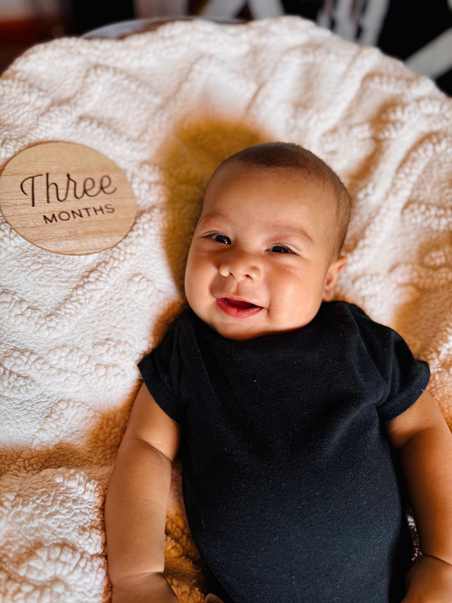 Noah is registered to the contest to win money with this photo: baby, infant, smile, black_onesie, blanket, wooden_plaque, text, three_months, face, skin, cute, lying_down, happy, young_child, portrait, indoors, soft_texture, warm_lighting, milestone, person