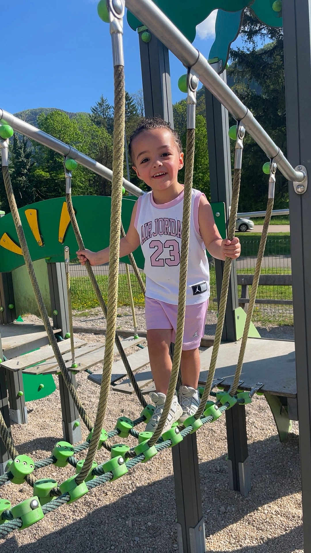 Nawël a rejoint le concours — aidez-le/la à gagner de superbes lots ! child, playground, rope_bridge, outdoor, smiling, daylight, sporty_clothing, air_jordan, shorts, sneakers, trees, mountains, rope, metal_bars, fence, gravel, bench, happy, person, summer
