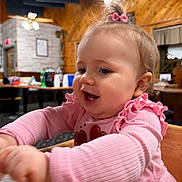 Laynee is registered to the contest to win money with this photo: baby, child, pink_clothing, bow, happy, laughing, indoor, wood_paneling, table, high_chair, long_sleeves, cute, person, infant, smiling, face, hands, restaurant, warm_lighting, casual