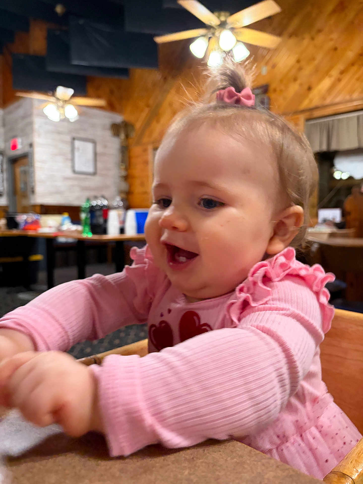 Laynee is registered to the contest to win money with this photo: baby, child, pink_clothing, bow, happy, laughing, indoor, wood_paneling, table, high_chair, long_sleeves, cute, person, infant, smiling, face, hands, restaurant, warm_lighting, casual