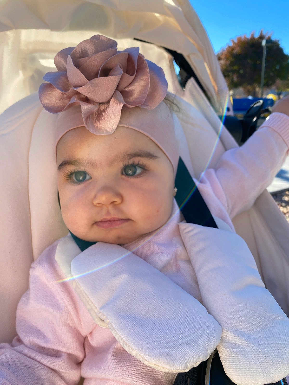 Leya is registered to the contest to win money with this photo: baby, child, stroller, headband, flower, pink_clothing, outdoor, sunlight, lens_flare, face, portrait, infant, cute, soft_tones, seatbelt, happy, daylight, person, young_child, closeup