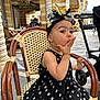 toddler, child, polka_dot_dress, headband, chair, wicker_chair, outdoor, side_view, gesture, baby, fashion, cute, portrait, stroller, columns, architecture, public_space, expression, seated, daylight