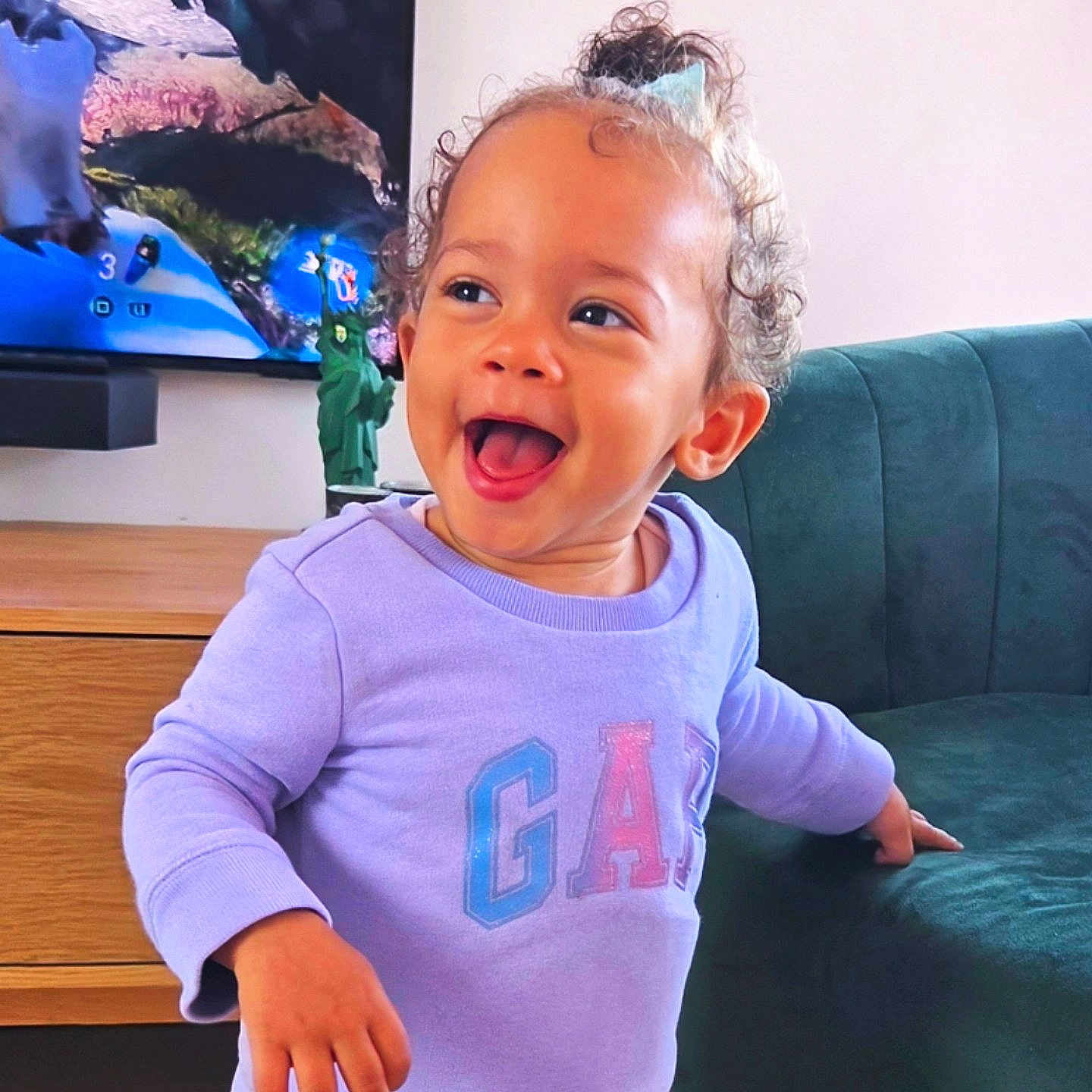 Arya participe au concours pour gagner de l'argent avec cette photo : toddler, child, smiling, curly_hair, purple_sweatshirt, gap_logo, living_room, couch, furniture, television, statue_of_liberty, toy, happy, indoors, person, young_child, hair_clip, wooden_furniture, playful, casual_clothing