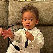 Arya a rejoint le concours — aidez-le/la à gagner de superbes lots ! toddler, child, pajamas, curly_hair, couch, headset, indoor, seated, cozy, expression, hand, face, person, furniture, home, curious, soft_lighting, textured, living_room, cute