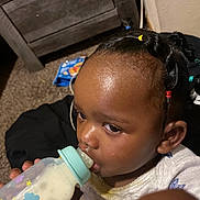 Ayla is registered to the contest to win money with this photo: baby_bottle, black_hair, cabinet, carpet, child, close_up, curious, curly_hair, cute, feeding, floor, furniture, hair_ties, indoors, milk, milk_bottle, person, toddler, white_clothing, young_child