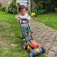 Ilyann participe au concours pour gagner de l'argent avec cette photo : child, toddler, toy, lawn_mower, grass, garden, pathway, greenery, outdoor, play, concentration, sweater, shorts, shoes, plants, fence, car, daylight, nature, yard