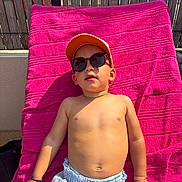 Ayden a rejoint le concours — aidez-le/la à gagner de superbes lots ! blue_shorts, bright, cap, casual, child, daytime, fence, leisure, outdoor, person, relaxing, shirtless, skin, summer, sunbathing, sunglasses, sunlight, towel, vacation, young