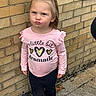 child, toddler, girl, portrait, standing, pouting, serious_expression, ponytail, pink_shirt, ruffles, hearts, black_pants, yellow_shoes, brick_wall, sidewalk, outdoor, daylight, fashion, full_body, humorous