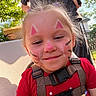 child, toddler, portrait, smiling, face_paint, outdoor, sunlight, backyard, red_shirt, baby_carrier, harness, seated, blonde_hair, purple_hair_ties, playful, happy, adult, trees, house, close_up