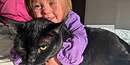 Elaina is registered to the contest to win money with this photo: child, black_cat, hugging, bed, purple_sweater, person, indoor, smiling, pet, feline, cozy, happy, casual_clothing, animal, human, portrait, resting, cute, family, comfort