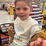 Jaxson is registered to the contest to win money with this photo: child, shopping_cart, grocery_store, toy, stuffed_animal, clothing, face, person, indoor, supermarket, shelf, bottle, drink, product, floor, tile, hand, shorts, expression, casual