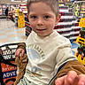 child, shopping_cart, grocery_store, boy, clothing, toy, hand, face, shorts, shelf, beverages, supermarket, casual, cute, indoor, person, store_aisle, smiling, young, retail