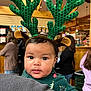 baby, child, reindeer_antlers, green, christmas_outfit, holiday, face, eyes, blanket, indoor, people, blurred_background, wood, warm_lighting, festive, person, cute, holding, headwear, expression