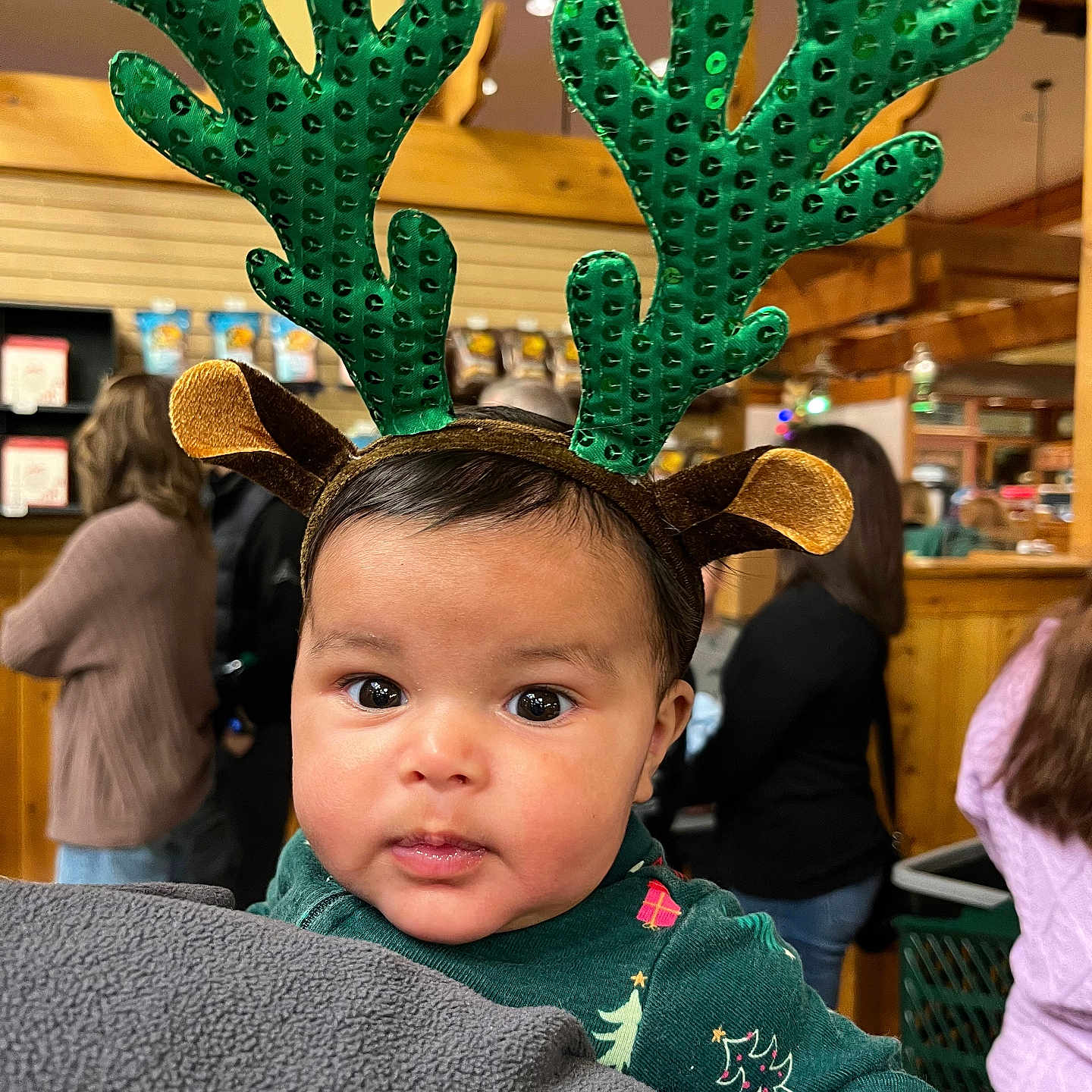 Hayden is registered to the contest to win money with this photo: baby, blanket, blurred_background, child, christmas_outfit, cute, expression, eyes, face, festive, green, headwear, holding, holiday, indoor, people, person, reindeer_antlers, warm_lighting, wood