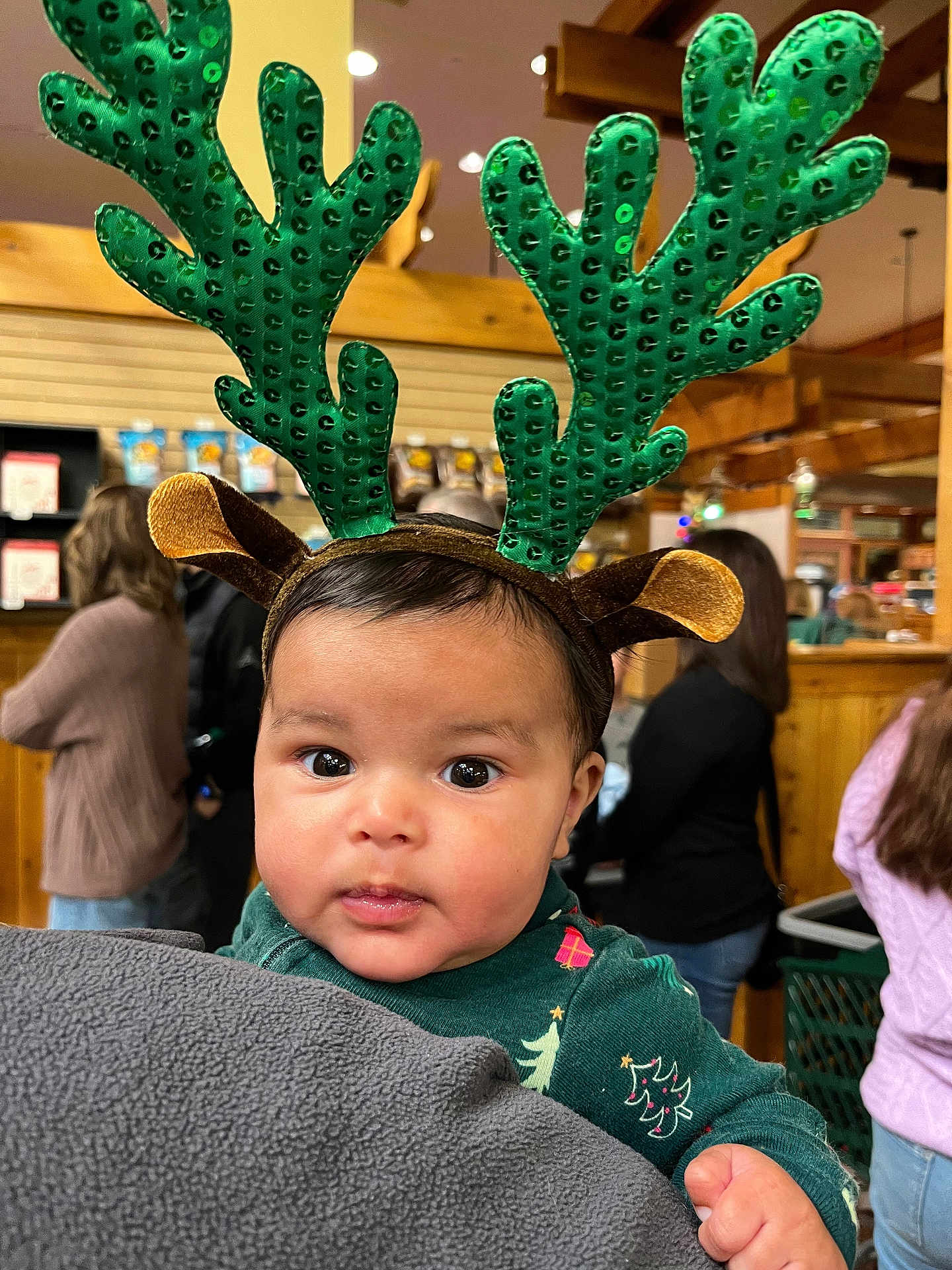 Hayden is registered to the contest to win money with this photo: baby, child, reindeer_antlers, green, christmas_outfit, holiday, face, eyes, blanket, indoor, people, blurred_background, wood, warm_lighting, festive, person, cute, holding, headwear, expression
