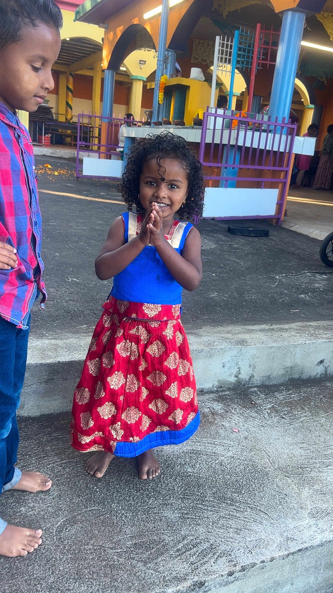 Maëllys a rejoint le concours — aidez-le/la à gagner de superbes lots ! child, girl, boy, traditional_dress, barefoot, curly_hair, smiling, outdoor, concrete, colorful, festival, cultural_event, pillar, purple_fence, casual_clothing, people, portrait, happy, daylight, hands_together