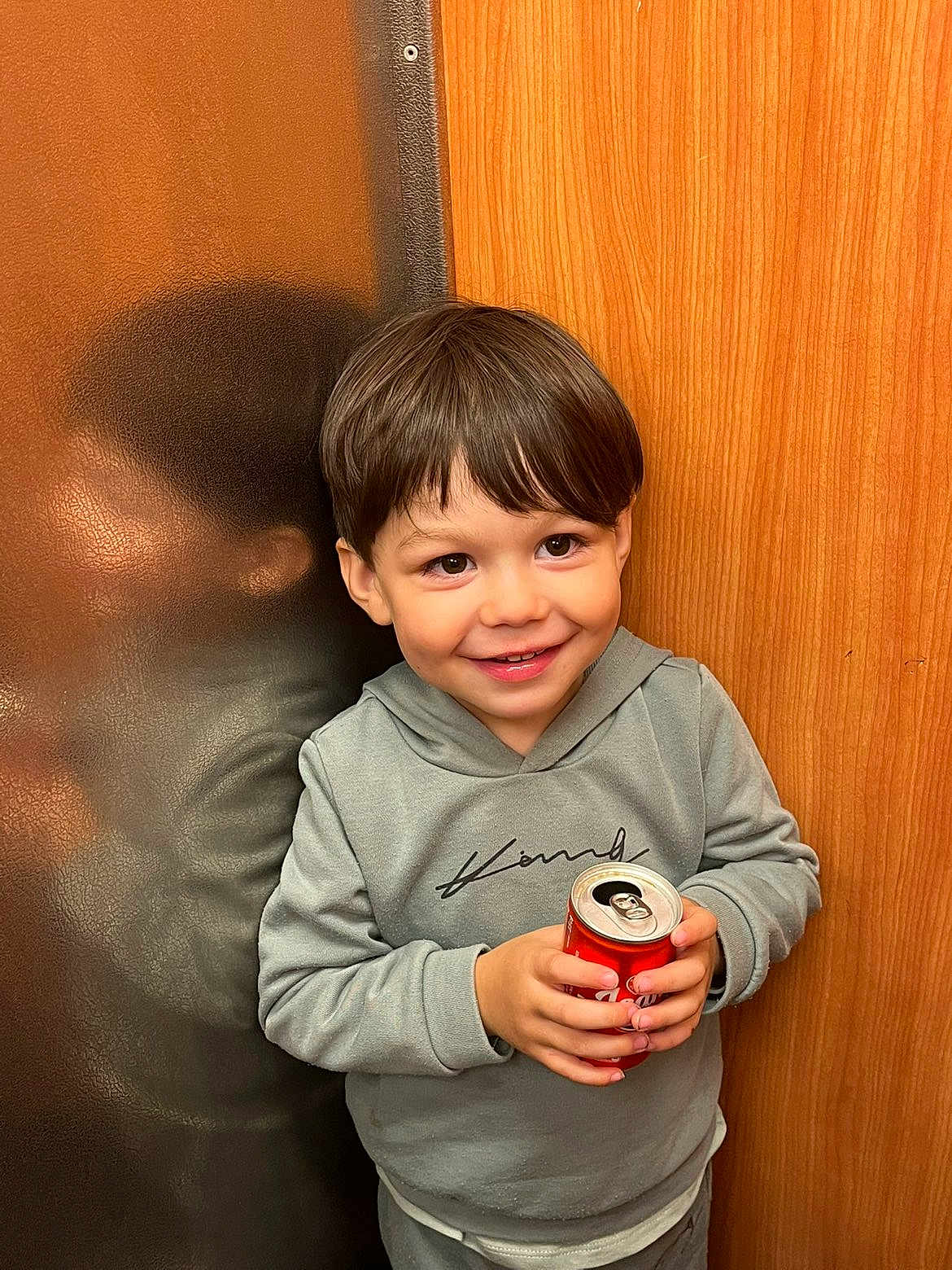 Imrân participe au concours pour gagner de l'argent avec cette photo : child, boy, smiling, soda_can, reflection, hoodie, short_hair, indoor, wood_panel, leather_surface, happy, casual_clothing, portrait, young, cute, holding_object, person, face, standing, bright