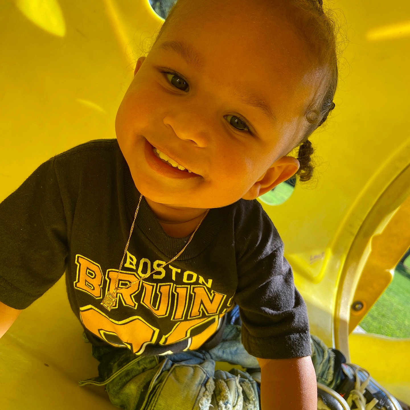 Zaelyn is registered to the contest to win money with this photo: black_tshirt, boston_bruins, braided_hair, casual_clothing, child, cute, happy, jeans, outdoor, person, play, playground, portrait, ripped_jeans, smiling, sneakers, sunlight, toddler, yellow_tunnel, young_child