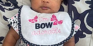 Lehlani is registered to the contest to win money with this photo: baby, infant, onesie, bib, mittens, patterned_clothing, lying_down, blanket, pillow, cute, face, hair_tie, indoor, child, adorable, pink, white, black, spot_pattern, expression