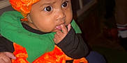 Lehlani joined the competition — help win amazing prizes! baby, child, pumpkin_costume, orange_hat, green_collar, cute, indoors, person, face, costume, holiday, halloween, plush, black_clothing, expression, finger_in_mouth, wide_eyes, sitting, legs, adult_hand